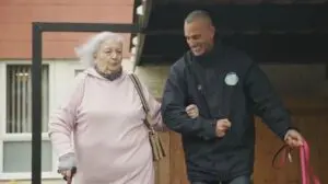 Elderly woman with walking stick being assisted by pet taxi driver while leaving her home for a pet appointment.