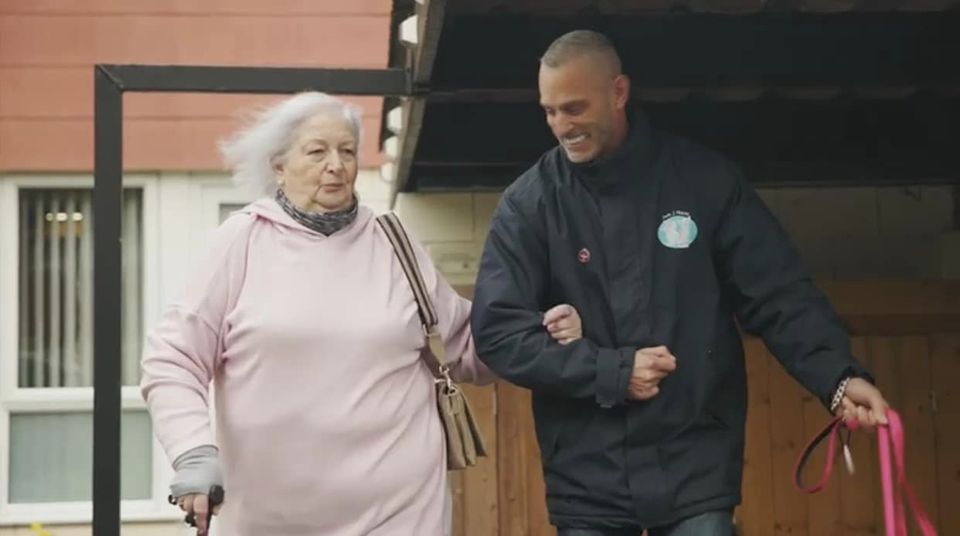 Elderly woman with walking stick being assisted by pet taxi driver while leaving her home for a pet appointment.